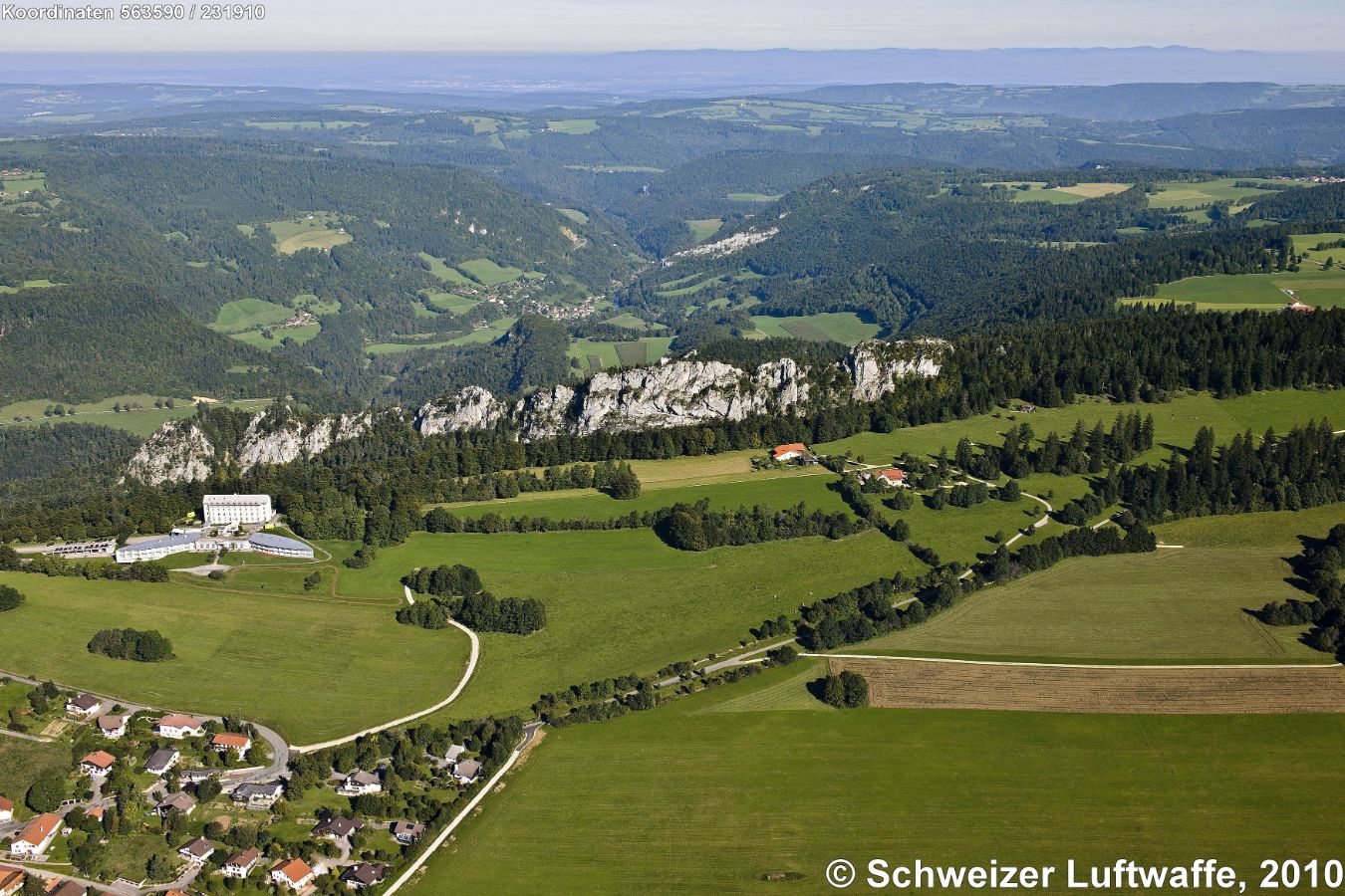 Arête de Sommêtres (Felswand Bildmitte). Siedlung vorne links: Le Noirmont. Grosses Gebäude Bildmitte links: Hôpital 'Clinique de Noirmont'. Siedlung im Talboden: Goumois (F). Im Hintergrund der Plateaujura.